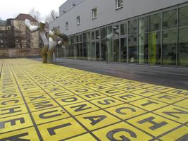 Random black letters on yellow background on the ground; metal sulpture of curvy pipes in background