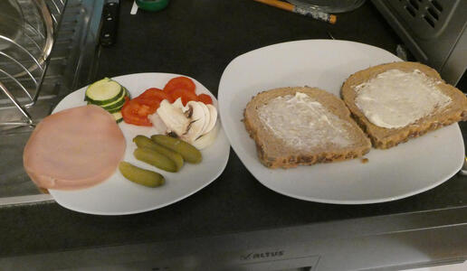 Two pieces of bread with mayonnaise on a plate. On another plate, two slices of bologna, pickles, zucchini, mushrooms, and tomatoes.