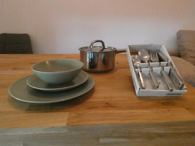 At left, a green plate, small plate, and bowl stacked up. In middle, a small steel pot. On right, a set of forks, knives, and spoons.