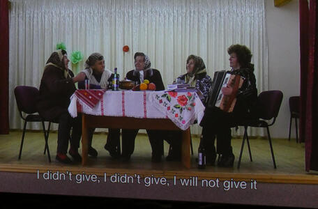 Women in Eastern European dress around a table; they are singing, and one is playing an accordion.
