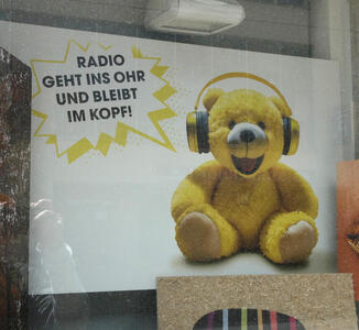 Yellow teddy bear wearing headphones. Text: Radio geht ins Ohr und bleibt im Kopf!