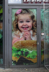 Door-sized photo of small girl with curly hair and pink flower in hair. She is smiling with glee and clasping her hands as she looks at a large hamburger.
