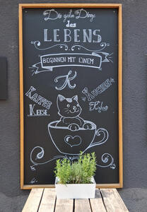 Chalkboard drawing of smiling cat in coffee cup. Text: „Die guten Dinge des Lebens beginnen mit einem K: Kaffee Kuchen Kakao Kekse“
