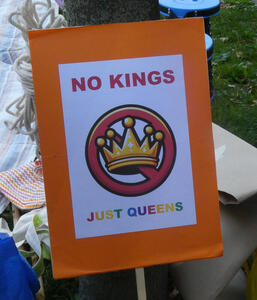 Sign: No Kings / Just Queens.  Second line is in rainbow colors. Has a picure of a crown with a red slash circle around it.