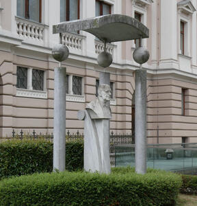 Bust of man wearing glasses. It is surrounded by short bushes and three stone columns with spheres at the top