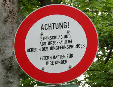 Red bordered circular sign with words: Achtung! Steinschlag und Absturzgefahr im Bereich des Jungfernsprunges. Eltern haften für Ihre Kinder