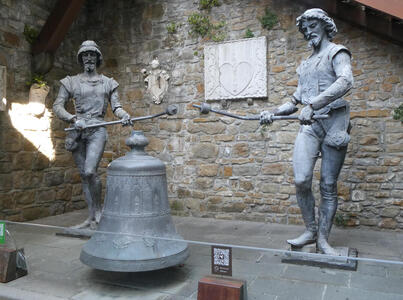 Statues of two bearded men holding staffs with hammer heads; they are used to strike a large bell in the foreground