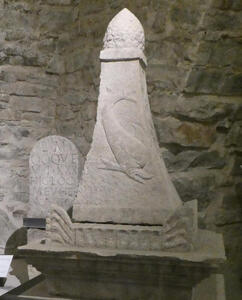 Pyramidal stone with sea creature in relief on side. To its left, a gravestone-shaped stone with a Latin inscription.