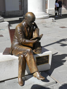 Bronze statue of a bald man seated, reading a book. There is a stack of several books next to him.