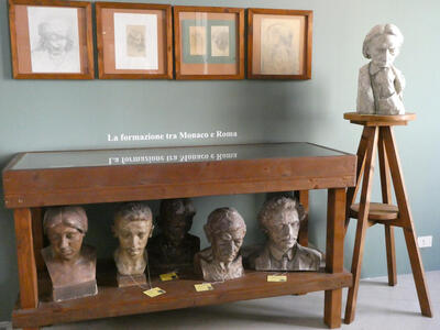 Five busts on a shelf under a table; a sixth bust of a man with chin resting on hand is atop a four-legged stool
