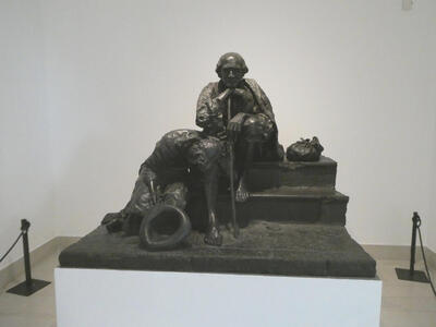 Statue of a seated man leaning on a walking stick. Next to him, a man bending down, his hat on the ground.