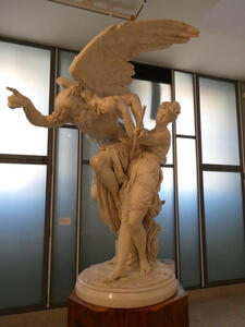 Statue of bearded man with wings, pointing forwards and turning his head towards a woman standing next to him.