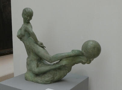 Sculpture of woman with hands behind her back. She is looking backwards towards her feet, where she is balancing a ball.