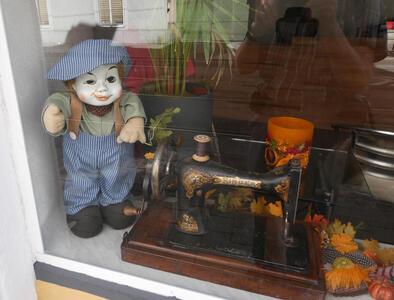 An antique sewing machine in a shop window. To its left, an antique doll in men’s work clothes.
