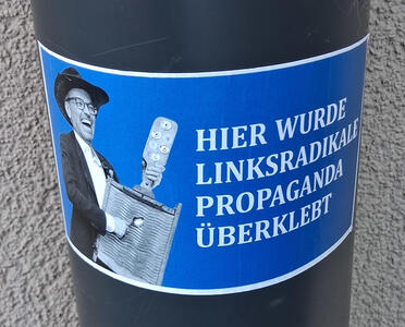 Sticker with Herbert Kickl, smiling, playing a washboard with a spoon. Text: HIER WURDE LINKSRADIKALE PROPAGANDA ÜBERKLEBT