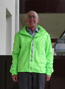 Bald man with glasses, wearing bright green rain jacket with reflective stripes. He is standing on a balcony of an apartment.