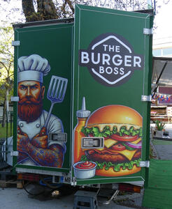 Artwork for “The Burger Boss” showing a large hamburger on one back door of a truck. On the other door is a glowering man with tattooed arms and a red beard and chef’s hat. He is holding a spatula.