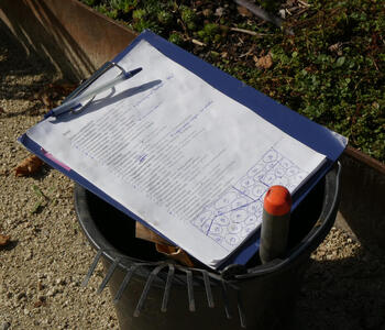Clipboard with a checklist of plants and a diagram of their locations. It is sitting on top of a bucket that has a trowel and a small rake in it.