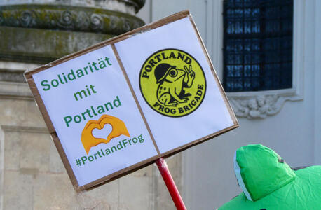 Sign with text “Solidarität mit Portland” and hands forming a heart. At right, a logo showing a frog wearing a helmet giving a V sign; text on logo is Portland Frog Brigade