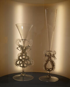 Two wine glasses, one extremely tall. Both have intricately designed stems with empty space in them.