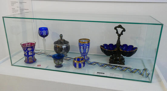 Display case with drdinking glasses, a goblet, a jar, a bowl for holding candies etc, and a long scepter-like object with spiral blue and yellow lines along its length.