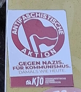 Circle with two flags; in circle is text Antifaschistische Aktion. Below circle: Gegen Nazis. Für Kommunismus. Damals wie heute.