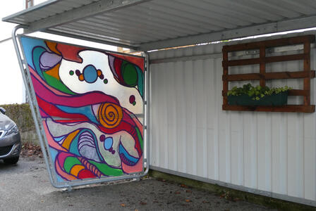 Bus shelter; one side has a colorful abstract painting. At the back of the shelter is a planter.