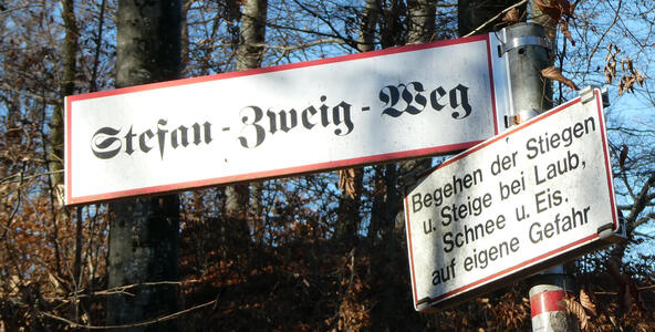 Street sign in Fraktur for Stefan-Zweig-Weg. A sign nearby says „Begehen der Stiegen u. Stiege bei Laub, Schnee u. Eis, auf eigene Gefahr“