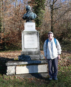Person in glasses, puffy white jacket, and English driving cap standing next to bust of Mozart