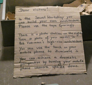 Dear visitors! In the Secret Workshop you can build your own architecture. Please use the tape sparingly. / There is a photo station on the right. Take a photo of your model in Rob Voerman’s high-rise architecture / You can use the torch on your mobile phone to illuminate it. / You can achieve a dramatic perspective by turning your mobile phone camera upside down.