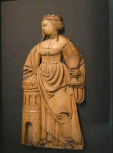 Wooden carving of Saint Barbara. She is rather plump, and she is standing next to a stone tower, which looks small in comparison to her. In one hand she has a wine goblet.