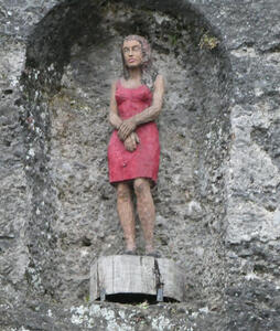 Niche with statue of woman in pink dress, arms folded across her waist.