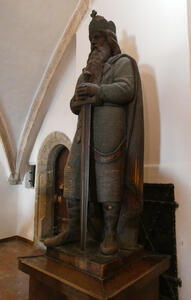 Bearded man in chain mail. He is wearing a crown with a cross on it and is holding a broadsword