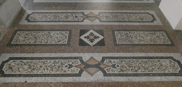 Geometric patterns in mosaic tile on floor. In center, a black square with embedded whie diamond and a four-way symmetric flower in brown and black.