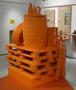 All-orange display of pallets, atop which are a barrel, a tire, and a small crate