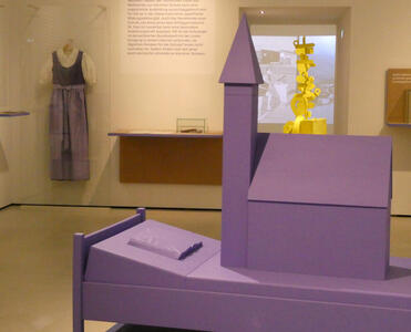 All-purple bed with a model of a church on top of it; in background, a purple dirndl