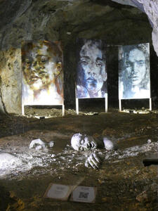 in background, three paintings of women’s faces. In foreground, a sculpture of a person’s head and hands, placed as if it is sinking into the ground.