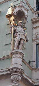 Statue of knight with gold helmet and spear; statues is on the corner of a building on an upper floor.