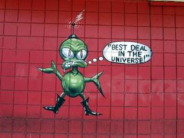 Green alien painted on wall of hat shop; thought balloon reads “Best deal in the universe!”