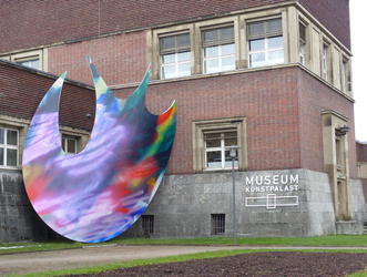 Near entrance of Museum Kunstpalast, very large irregulary shaped colored art piece leaning on building.