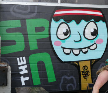 Cartoon face in light bluewith headband in Austrian flag colors (red/whie/red horizontal stripes). The eyes form the “o”s in the text “the spoon”