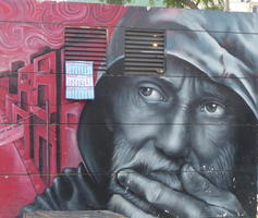Bearded man in gray, hand on chin, looking at red buildings in background