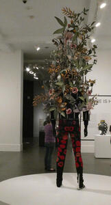 Statue of person in black and red pants, with flowers and branches growing out of back.