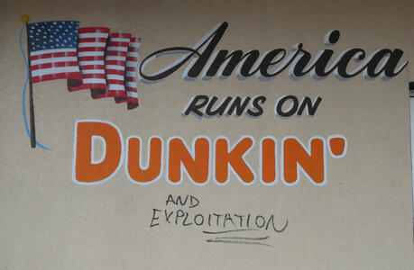 Sign: America runs on Dunkin’; underneath someone has written in marker “and exploitation”