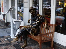 Bronze sculpture of shoeless man in hat, seated on a wooden bench