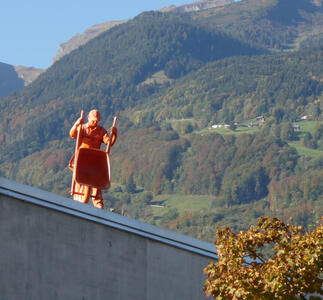 Large orange sculpture of a worker in a safety helmet. He is holding a wheelbarrow vertically, as if emptying its contents. Forested mountain in background