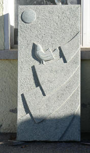 Rectangular gray stone. At top, a circle representing the sun. In middle, a bird perched on roof.