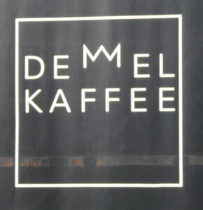 Logo for Demmel Kaffee; the two Ms are joined to make a crown and are positioned above the other letters.