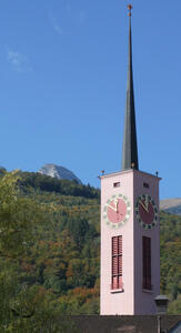 Church tower. Two sides have a clock with a pink face. Mountain in background.