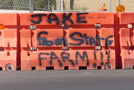 Orange traffic barrier with “Jake from State Farm!!!” spray-painted on it